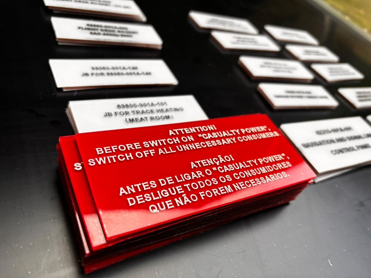 Placas de identificação em acrílico vermelho com letras brancas gravadas e placas brancas com letras pretas, produzidas pela Grava CNC